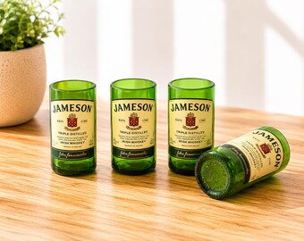 May include: Four drinking glasses crafted from green glass Jameson Irish Whiskey bottles. Three glasses stand upright, while one lies on its side. Each glass features the cream-colored Jameson label with black text and the red crest logo.