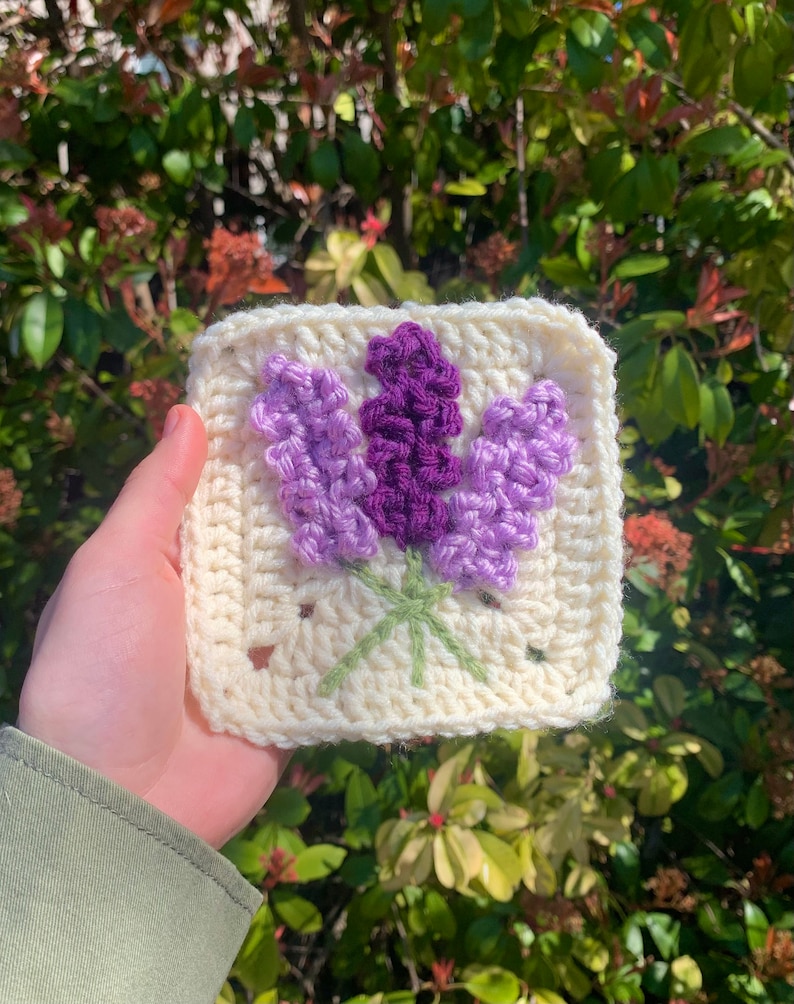 May include: A crocheted square with a lavender flower design. The flower is made with purple and light purple yarn, and the stems are green. The square is made with white yarn.