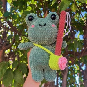 Puede incluir: Una muñeca de rana de ganchillo hecha a mano en tonos verdes, con ojos negros y mejillas rosas. Tiene una correa amarilla y un accesorio rojo y rosa. La rana es suave y afelpada, ideal para un juguete infantil.
