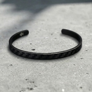 Op de afbeelding: Een zwarte manchetarmband met een koolstofvezelpatroon. De open armband is gemaakt van donker metaal en heeft een subtiel, gestructureerd ontwerp. De armband wordt getoond op een grijze ondergrond.