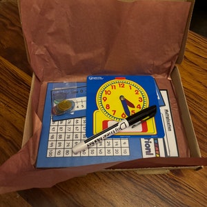 Può includere: Una scatola di cartone piena di risorse educative per bambini. La scatola contiene un orologio blu e giallo, un pennarello cancellabile a secco, una tabella di moltiplicazione e una tabella di linea numerica. La scatola è aperta e il contenuto è visibile.