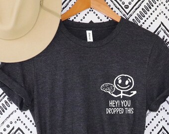 May include: Dark grey t-shirt featuring a white graphic of a stick figure with a brain and the text "HEY! YOU DROPPED THIS." A beige fedora hat hangs above the shirt on a wooden hanger. The background has a black and white zigzag pattern.