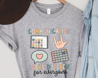 Puede incluir: Camiseta gris con la frase "COMMUNICATION looks DIFFERENT for everyone" en una fuente colorida. La camiseta presenta ilustraciones de una tableta, una mano haciendo el signo "Te amo", una calculadora y un bocadillo.