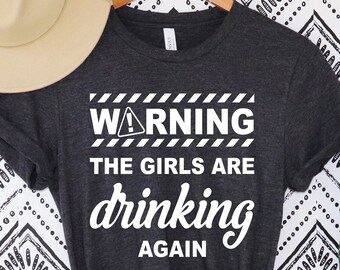 Pode incluir: Camiseta cinza escuro com o texto "WARNING THE GIRLS ARE drinking AGAIN" em branco. Um chapéu bege repousa no canto superior esquerdo. A camisa está pendurada em um cabide de madeira. Uma planta verde alta está à direita.