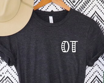 May include: Dark gray t-shirt with the white letters "OT" on the front. A beige hat hangs above the shirt on a wooden hanger. A tall green plant in a white pot is to the right. The background is a black and white zigzag pattern.