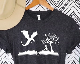 May include: Dark gray t-shirt featuring a white graphic of a dragon, an open book, a tree, and a person reading. The shirt is displayed on a wooden hanger with a beige hat in the background. The shirt is knotted at the waist.