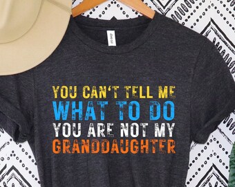 May include: Dark gray t-shirt with the text "YOU CAN'T TELL ME WHAT TO DO YOU ARE NOT MY GRANDDAUGHTER" in blue, orange, and yellow. A beige hat hangs above the shirt on a wooden hanger. The background is a black and white zigzag pattern.