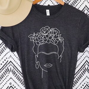 May include: A dark gray t-shirt featuring a white line drawing of a woman's face with a floral crown. A beige fedora hat rests above the shirt, and a tall green plant is to the right. The shirt is knotted at the waist.