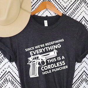 May include: Dark gray t-shirt with the text "SINCE WE'RE REDEFINING EVERYTHING THIS IS A CORDLESS HOLE PUNCHER" and a graphic of a handgun. A beige fedora hat rests on the top left. The background is a black and white zigzag pattern.