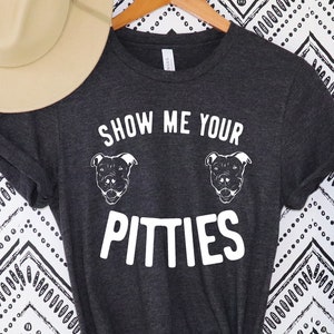 May include: Dark gray t-shirt with the text "SHOW ME YOUR PITTIES" in white, along with two dog illustrations. A tan hat rests above the shirt, and a green plant is to the right. The shirt is hanging on a wooden hanger.