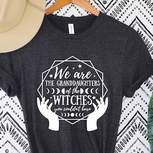 Peut inclure: T-shirt gris fonc&eacute; avec l'inscription blanche "We are the granddaughters of the witches you couldn't burn" dans un motif g&eacute;om&eacute;trique. Un chapeau beige est suspendu au-dessus du t-shirt sur un cintre en bois.