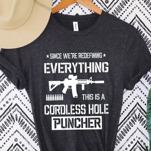 May include: Dark gray t-shirt with the text "SINCE WE'RE REDEFINING EVERYTHING THIS IS A CORDLESS HOLE PUNCHER" in white. The design includes a graphic of a rifle and bullets. A beige hat hangs above the shirt.