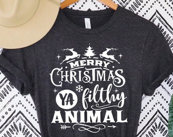 Könnte beinhalten: Dunkelgraues T-Shirt mit dem weißen Text "Merry Christmas Ya Filthy Animal". Das Design umfasst Rentiere, einen Weihnachtsbaum und Pfeile. Ein beiger Hut liegt oben links. Das Shirt hängt an einem Holzbügel.