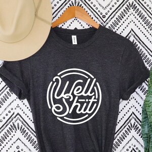 May include: Charcoal grey t-shirt featuring the white text "Well Shit" within a circular design. A beige hat is positioned above the shirt, with a potted snake plant to the right. The shirt hangs on a wooden hanger against a monochrome patterned backdrop.