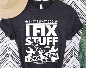 May include: Dark gray t-shirt with the text "THAT'S WHAT I DO, I FIX STUFF AND I KNOW THINGS" in white. The design includes wrenches and a hand holding a wrench. A beige hat hangs above the shirt.