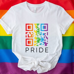 May include: White t-shirt featuring a colorful QR code design in rainbow colors. The word "PRIDE" is printed in black below the QR code. The shirt is displayed on a rainbow flag background.