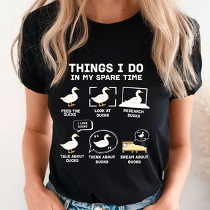 K&ouml;nnte beinhalten: Schwarzes T-Shirt mit dem Text "THINGS I DO IN MY SPARE TIME" und Enten-Illustrationen. Das Design zeigt Aktivit&auml;ten wie F&uuml;ttern, Betrachten und Recherchieren von Enten sowie "I LOVE DUCKS".