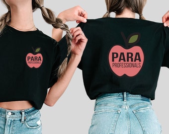 May include: Black t-shirts featuring a red apple graphic with the words "PARA PROFESSIONALS" in white. The apple design is on the front and back of the shirts. The shirts are being held up by a person.