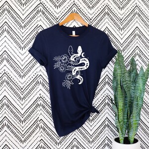 May include: Navy blue t-shirt featuring a white graphic of two snakes intertwined with floral and moon elements. The short-sleeved shirt is hanging on a wooden hanger against a patterned background.