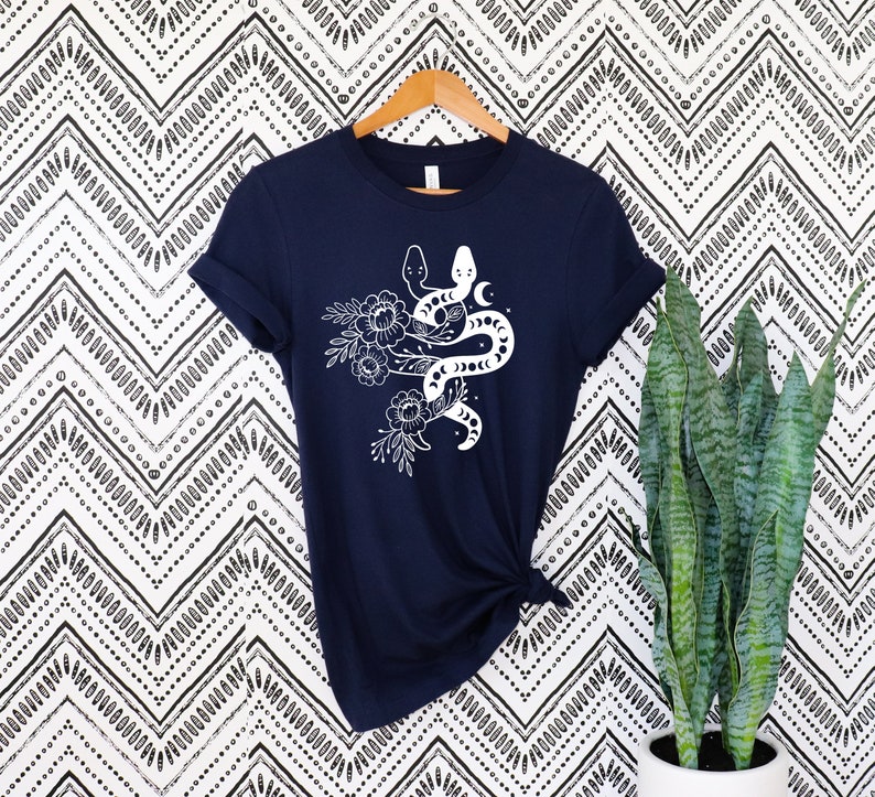 May include: Navy blue t-shirt featuring a white graphic of two snakes intertwined with floral and moon elements. The short-sleeved shirt is hanging on a wooden hanger against a patterned background.