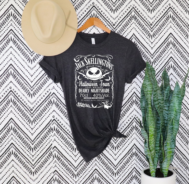 May include: Dark gray t-shirt with a white graphic featuring Jack Skellington from The Nightmare Before Christmas. The graphic includes text that reads "Jack Skellington's Mold Time Halloween Town Deadly Nightshade 70cl 40%Vol."