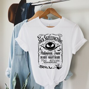 May include: White t-shirt featuring a black graphic with the text "Jack Skellingtons" and "Halloween Town". The design includes a stylized image of Jack Skellington's face, bats, and other Halloween-themed elements. The shirt is hanging on a wooden hanger.