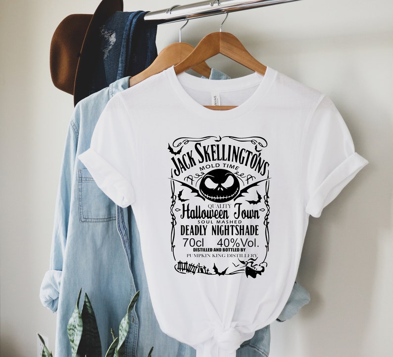 May include: White t-shirt featuring a black graphic with the text "Jack Skellingtons" and "Halloween Town". The design includes a stylized image of Jack Skellington's face, bats, and other Halloween-themed elements. The shirt is hanging on a wooden hanger.