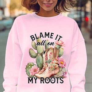 Op de afbeelding: Een lichtroze sweatshirt met een afbeelding van cowboylaarzen, cactussen en bloemen. De tekst "BLAME IT all on MY ROOTS" is boven de afbeelding gedrukt. De sweatshirt wordt door een persoon gedragen.
