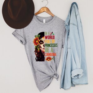 Pode incluir: Uma camiseta cinza com um desenho de caveira de a&ccedil;&uacute;car com flores e o texto "IN A WORLD FULL OF PRINCESSES BE A CHINGONA". Um chap&eacute;u marrom e uma jaqueta jeans tamb&eacute;m est&atilde;o presentes.