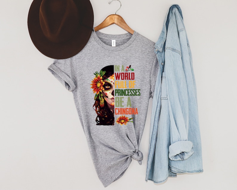Pode incluir: Uma camiseta cinza com um desenho de caveira de a&ccedil;&uacute;car com flores e o texto "IN A WORLD FULL OF PRINCESSES BE A CHINGONA". Um chap&eacute;u marrom e uma jaqueta jeans tamb&eacute;m est&atilde;o presentes.