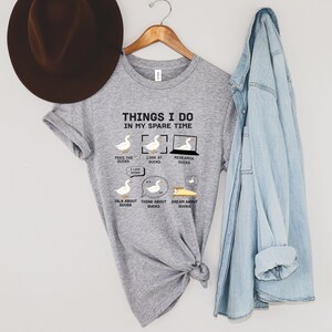 K&ouml;nnte beinhalten: Ein hellgraues T-Shirt mit dem Text "THINGS I DO IN MY SPARE TIME" und Entenillustrationen. Das Shirt zeigt Enten bei verschiedenen Aktivit&auml;ten, wie F&uuml;ttern, Betrachten und Recherchieren von Enten.
