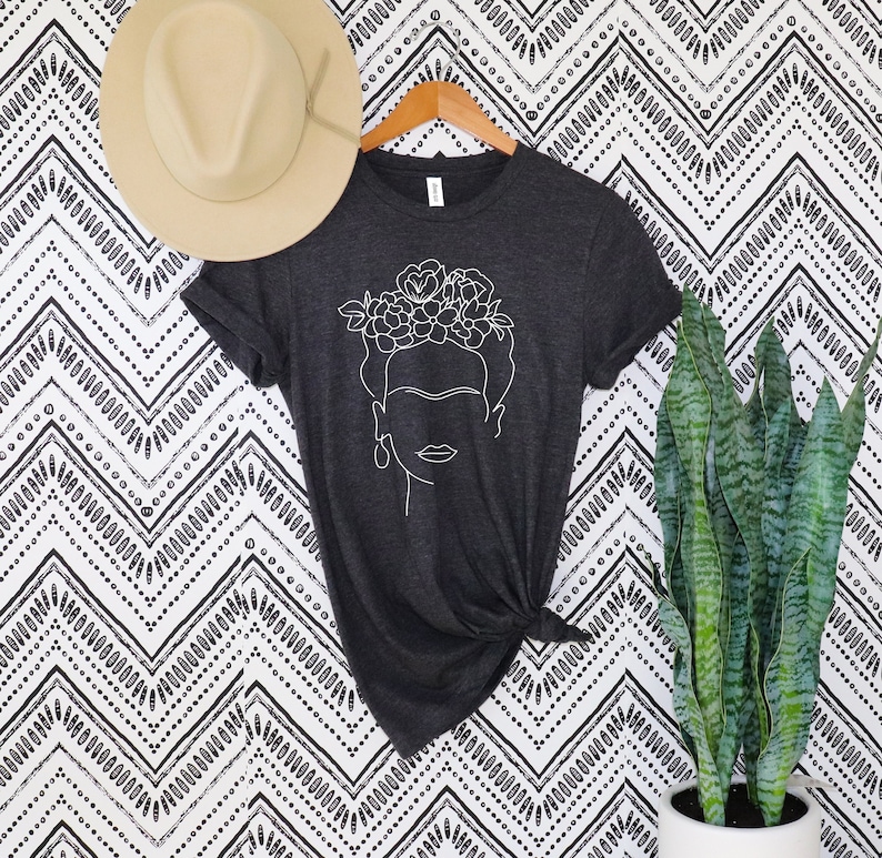 May include: A dark gray t-shirt featuring a white line drawing of a woman's face with a floral crown. A beige fedora hat rests above the shirt, and a tall green plant is to the right. The shirt is knotted at the waist.