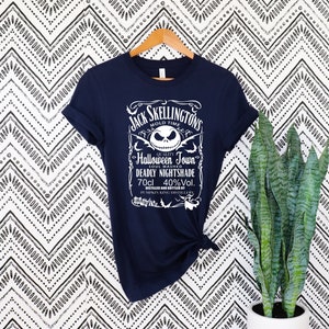 May include: Navy blue t-shirt featuring a white graphic with the text "Jack Skellington's Halloween Town Deadly Nightshade." The design includes a skull and other decorative elements. The shirt is hanging on a wooden hanger.