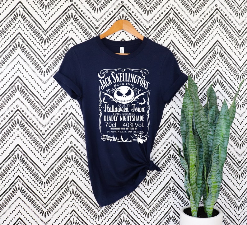 May include: Navy blue t-shirt featuring a white graphic with the text "Jack Skellington's Halloween Town Deadly Nightshade." The design includes a skull and other decorative elements. The shirt is hanging on a wooden hanger.