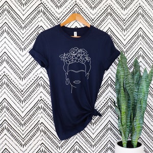 May include: Navy blue t-shirt featuring a white line drawing of a woman with a floral crown and earrings. The shirt is hanging on a wooden hanger against a black and white patterned background. The shirt is knotted at the waist.