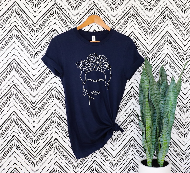 May include: Navy blue t-shirt featuring a white line drawing of a woman with a floral crown and earrings. The shirt is hanging on a wooden hanger against a black and white patterned background. The shirt is knotted at the waist.