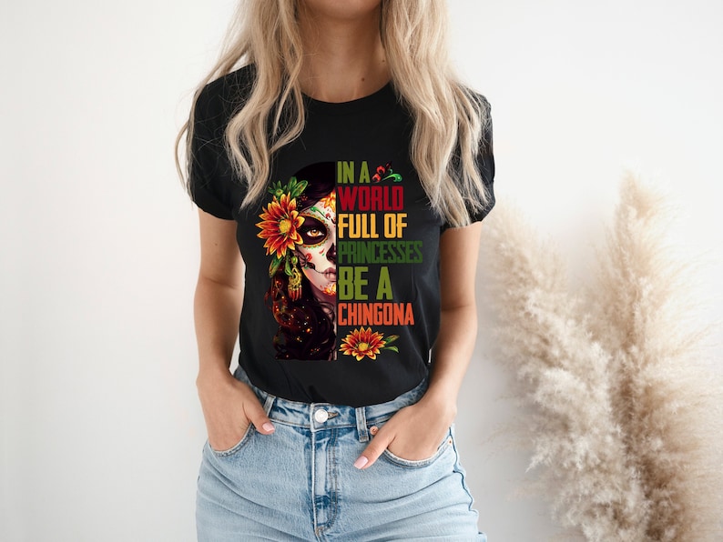 Pode incluir: T-shirt preto com um gr&aacute;fico colorido de caveira de a&ccedil;&uacute;car com detalhes florais e o texto "IN A WORLD FULL OF PRINCESSES BE A CHINGONA". O design inclui cores vermelhas, amarelas e verdes.