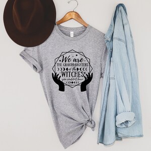 Peut inclure: T-shirt gris chin&eacute; avec l'inscription noire "We are the granddaughters of the witches you couldn't burn". Le motif comprend des mains, des lunes et des &eacute;toiles. Un chapeau marron et une chemise en jean sont &agrave; proximit&eacute;.