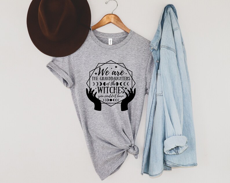 Peut inclure: T-shirt gris chin&eacute; avec l'inscription noire "We are the granddaughters of the witches you couldn't burn". Le motif comprend des mains, des lunes et des &eacute;toiles. Un chapeau marron et une chemise en jean sont &agrave; proximit&eacute;.