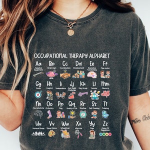 May include: Dark grey t-shirt featuring the text "OCCUPATIONAL THERAPY ALPHABET" and an alphabet chart with occupational therapy terms and illustrations. The shirt has a crew neck.