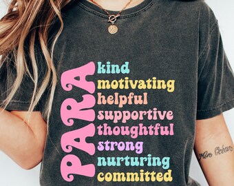 May include: Dark grey t-shirt featuring the word "PARA" in pink, and a list of positive attributes in various colours: kind, motivating, helpful, supportive, thoughtful, strong, nurturing, and committed. The shirt is worn by a person.