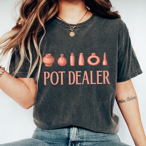 Puede incluir: Camiseta gris oscuro con el texto rosa "POT DEALER" sobre una fila de ilustraciones de cerámica rosa. Un estilo de top casual y cómodo.