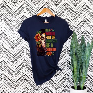 Pode incluir: Camiseta azul marinho com um gr&aacute;fico colorido de uma mulher com maquiagem de caveira de a&ccedil;&uacute;car, adornada com flores. O texto diz: "In a world full of princesses be a chingona". A camiseta est&aacute; pendurada em um cabide de madeira.
