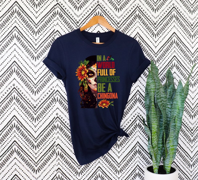Pode incluir: Camiseta azul marinho com um gr&aacute;fico colorido de uma mulher com maquiagem de caveira de a&ccedil;&uacute;car, adornada com flores. O texto diz: "In a world full of princesses be a chingona". A camiseta est&aacute; pendurada em um cabide de madeira.
