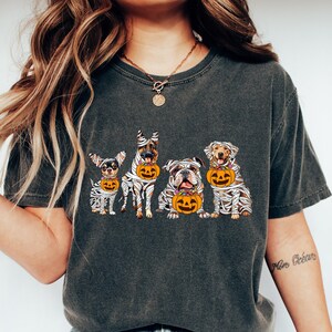 May include: Dark gray t-shirt featuring a Halloween design with five dogs dressed as mummies, each holding a jack-o'-lantern bucket. The dogs are different breeds, including a Chihuahua, German Shepherd, Bulldog, and Labrador Retriever.