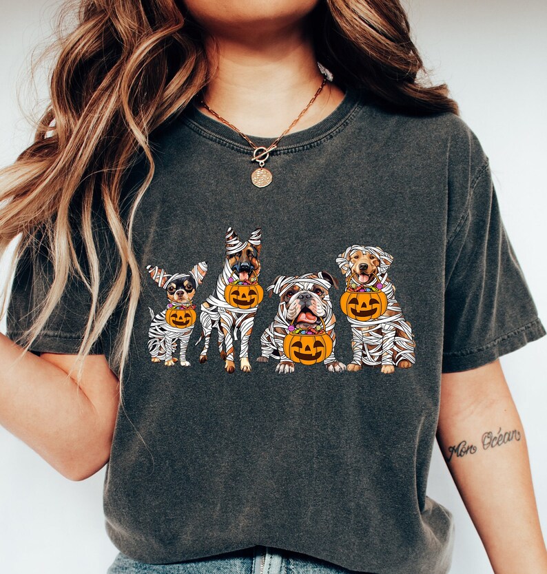 May include: Dark gray t-shirt featuring a Halloween design with five dogs dressed as mummies, each holding a jack-o'-lantern bucket. The dogs are different breeds, including a Chihuahua, German Shepherd, Bulldog, and Labrador Retriever.