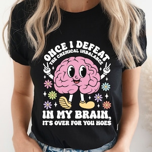 Puede incluir: Camiseta negra con un diseño de cerebro de dibujos animados. El cerebro tiene una cara sonriente, brazos y piernas, rodeado de flores. El texto dice: "ONCE I DEFEAT THE CHEMICAL IMBALANCE IN MY BRAIN, IT'S OVER FOR YOU HOES."
