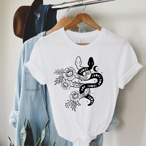 May include: White t-shirt featuring a black graphic of two snakes intertwined with floral elements and a crescent moon. The design is printed on a classic crew neck tee, perfect for casual wear.