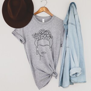 May include: A heather gray t-shirt featuring a black line drawing of a woman with a floral crown. The t-shirt is hanging on a wooden hanger, with a brown hat and a light blue denim jacket nearby. The shirt is knotted at the bottom.