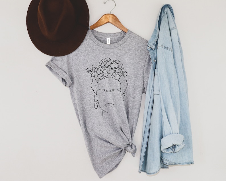 May include: A heather gray t-shirt featuring a black line drawing of a woman with a floral crown. The t-shirt is hanging on a wooden hanger, with a brown hat and a light blue denim jacket nearby. The shirt is knotted at the bottom.